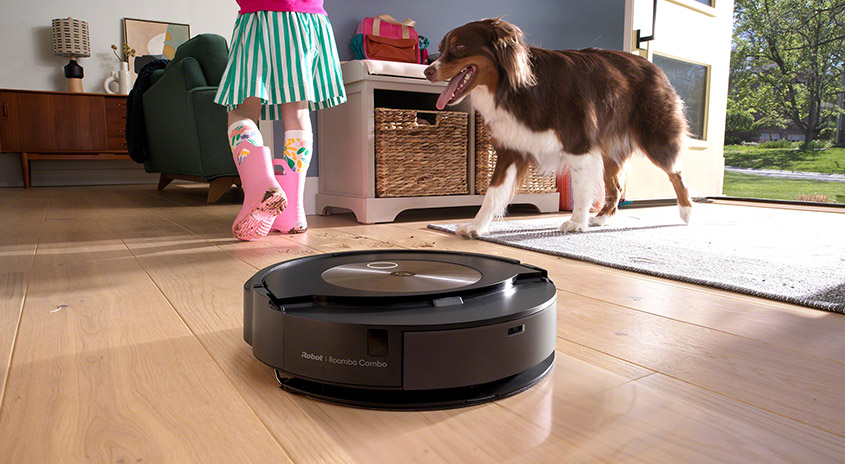 Ein schwarzer Roboterstaubsauger auf einem Holzboden, ein Hund und Kind im Hintergrund.