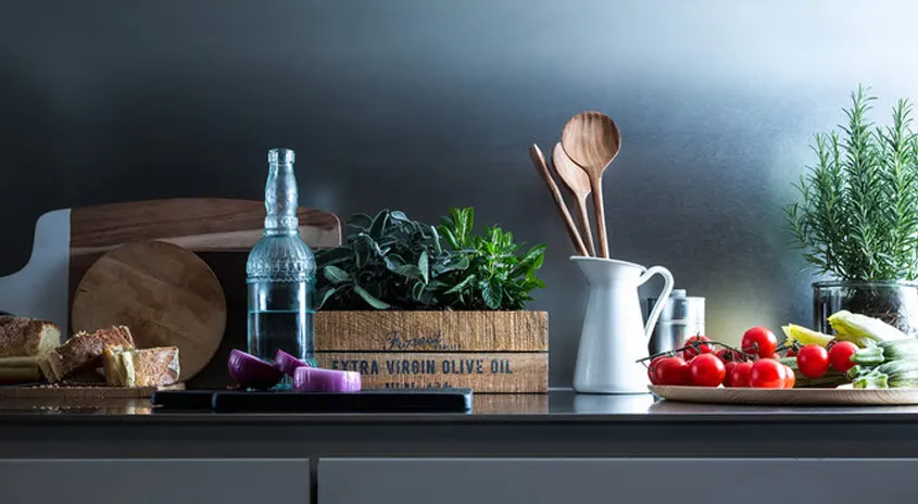 Auf einer Küchentheke stehen Lebensmittel, Kräuter, eine Flasche, eine Kanne und hölzerne Küchenutensilien vor einer grauen Wand.