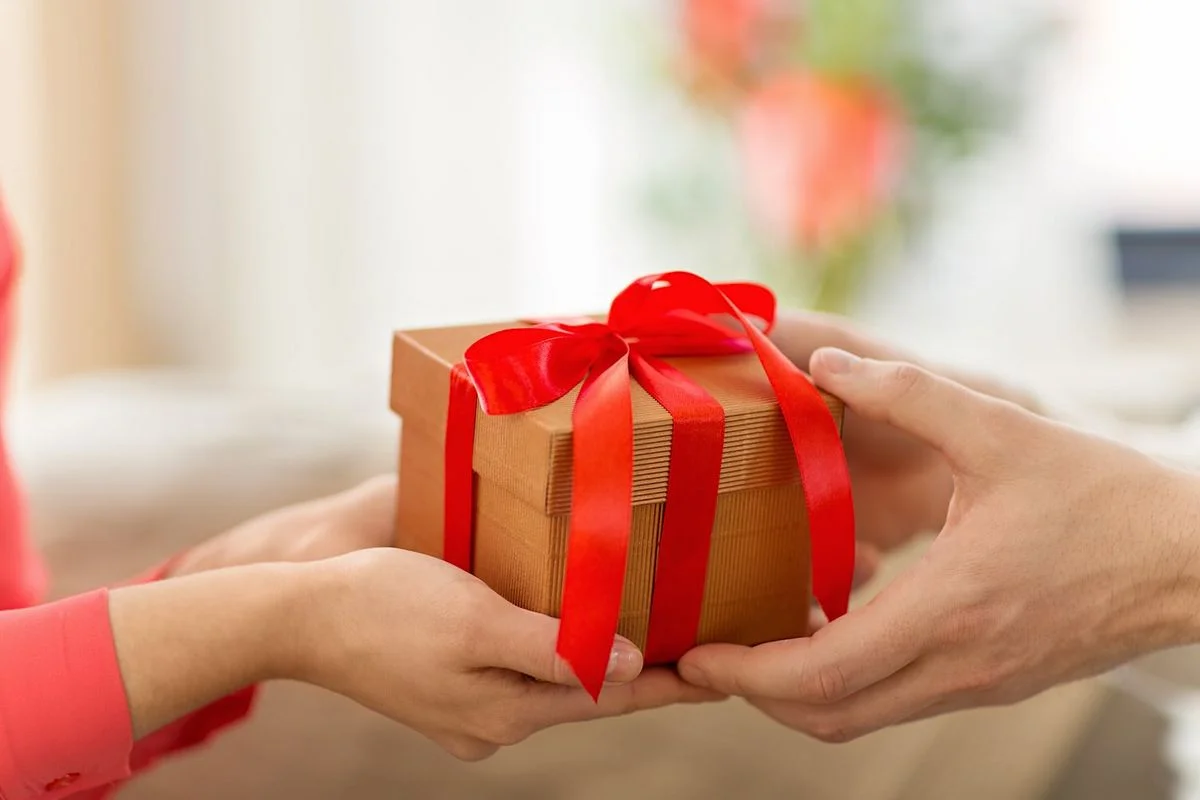 Zwei Personen halten gemeinsam eine kleine Geschenkbox aus Karton mit roter Schleife, die von einer Hand zur anderen überreicht wird.