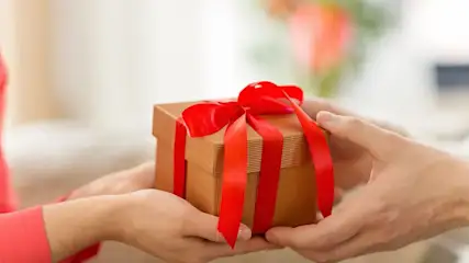 Zwei Personen halten gemeinsam eine kleine Geschenkbox aus Karton mit roter Schleife, die von einer Hand zur anderen überreicht wird.