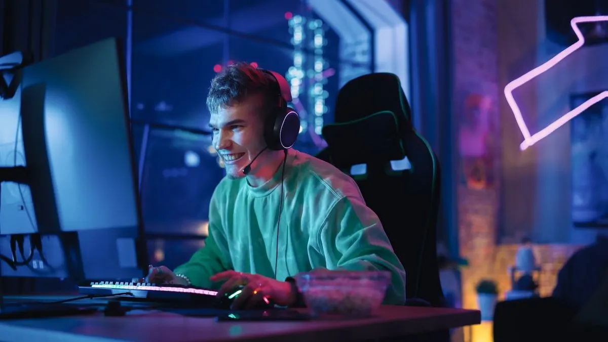 Lächelnder Mann in grünem Shirt mit Headset spielt an einem Computer mit beleuchteter Tastatur. Neonlicht und Snacks im Raum.