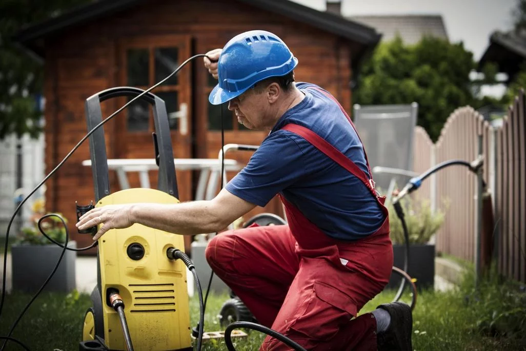 Ein Mann in roter Arbeitshose und Blauem Helm hantiert mit einem gelben Hochdruckreiniger