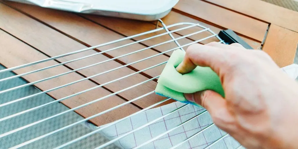 Eine Hand reinigt einen Grillrost mit einem gruene Schwamm auf einem Holztisch