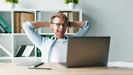 Lachender Mann mit Brille, Hände hinter dem Kopf, sitzt vor einem Laptop an einem Schreibtisch. Im Hintergrund weiße Regale mit Büroartikeln und Topfpflanzen.