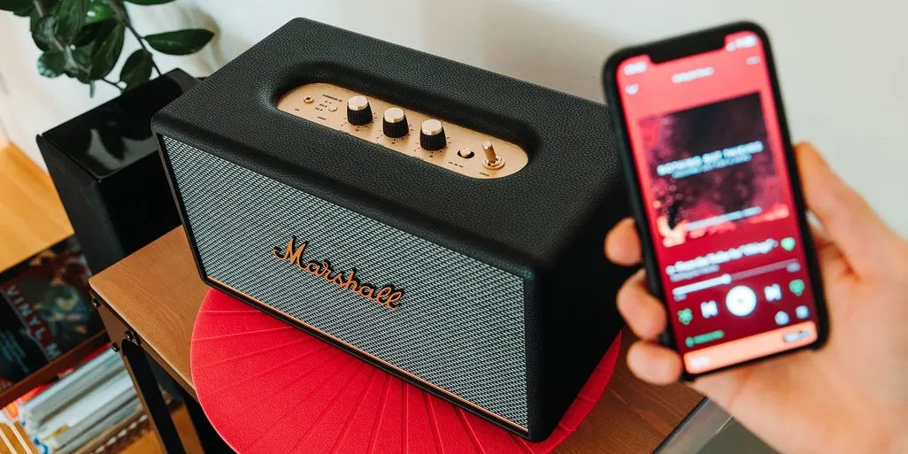 Schwarzer Marshall-Lautsprecher mit goldenem Bedienfeld und Logo auf roter Matte. Eine Hand hält ein Smartphone mit roter Musik-App.
