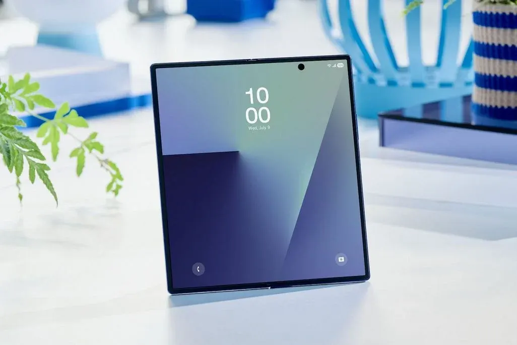 Samsung Galaxy Z Fold 7 aufgeklappt stehend; großes Innen‑Display zeigt Uhrzeit 10:00, mittig oben Lochkamera; Tisch mit Pflanzen und blauer Deko im Hintergrund.