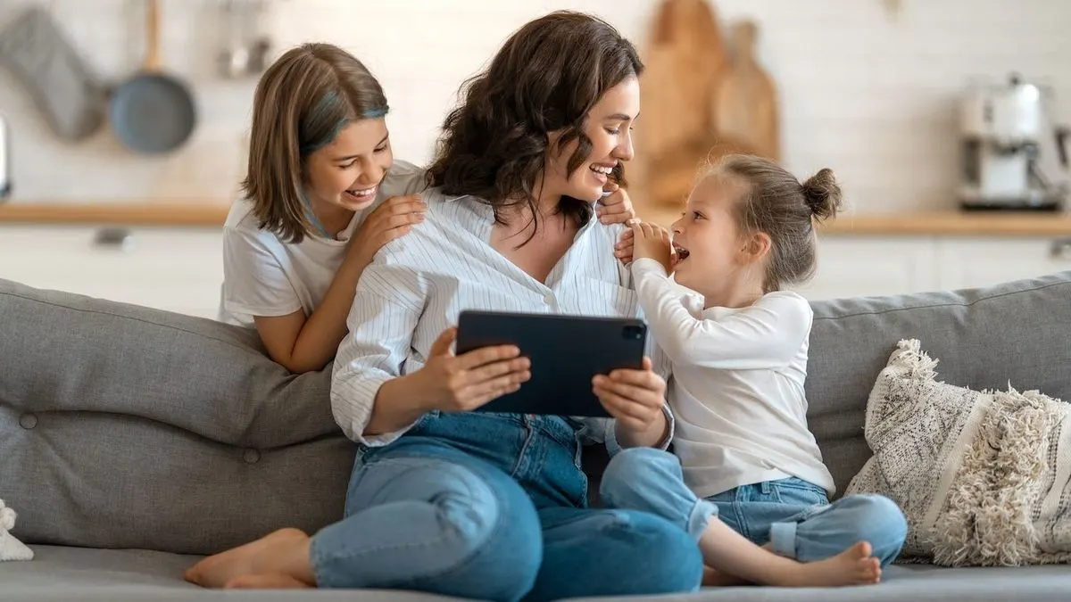 Lachende Frau mit zwei Kindern auf einem grauen Sofa, die ein schwarzes Tablet hält. Im Hintergrund ist eine moderne Küche zu sehen.