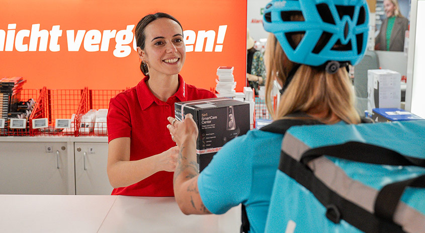 Ein Lieferant in blauem Hemd hält drei braune Kartons in einem Geschäft mit Regalen und einem Computer.