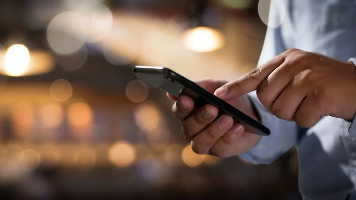 Hände in hellblauem Hemd halten ein schwarzes Smartphone und tippen auf den Bildschirm.