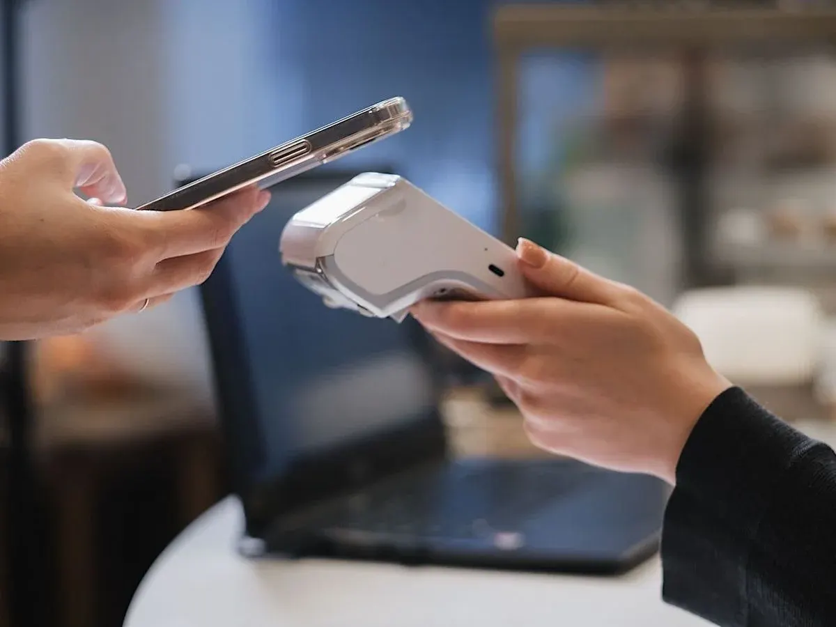 Kontaktlose Kartenzahlung: Person hält Smartphone an mobiles Kartenlesegerät vor aufgeklapptem Laptop.