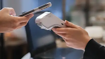 Kontaktlose Kartenzahlung: Person hält Smartphone an mobiles Kartenlesegerät vor aufgeklapptem Laptop.