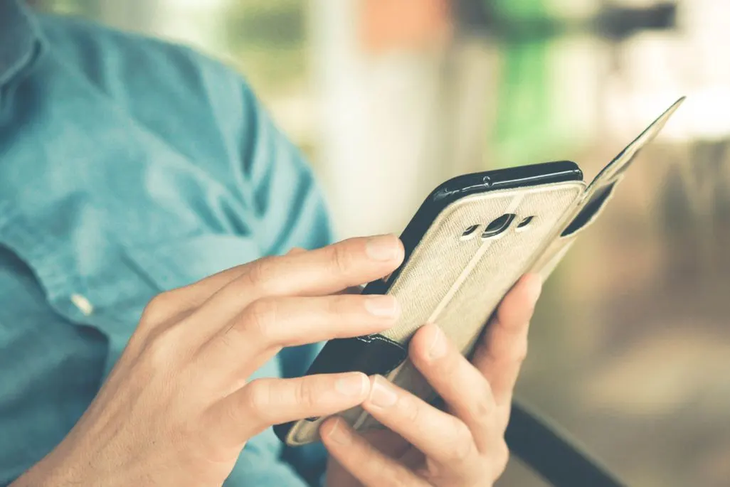 Eine Person in blauem Hemd hält ein Smartphone in beiger, texturierter Hülle. Der Hintergrund ist unscharf.