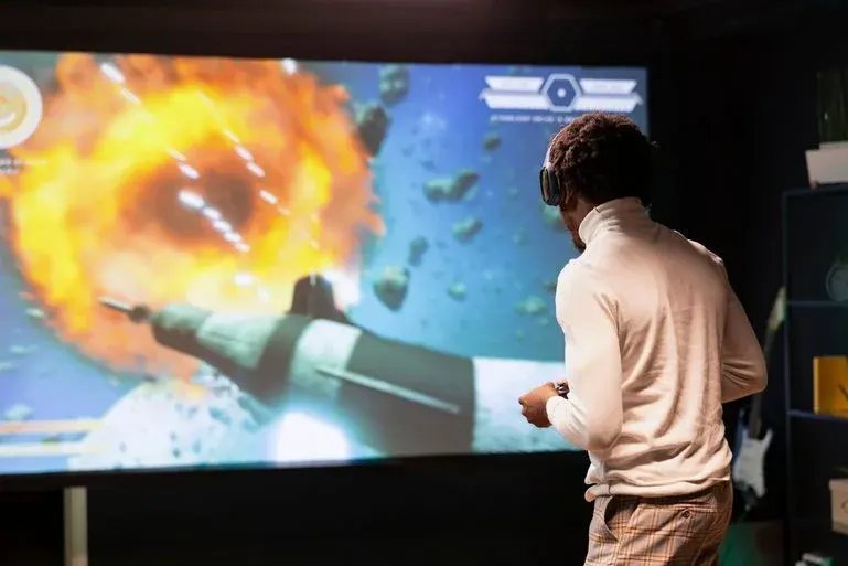 Person in weißem Rollkragenpullover und VR-Headset spielt mit Controller. Großer Bildschirm zeigt Weltraumexplosion und Raumschiff.