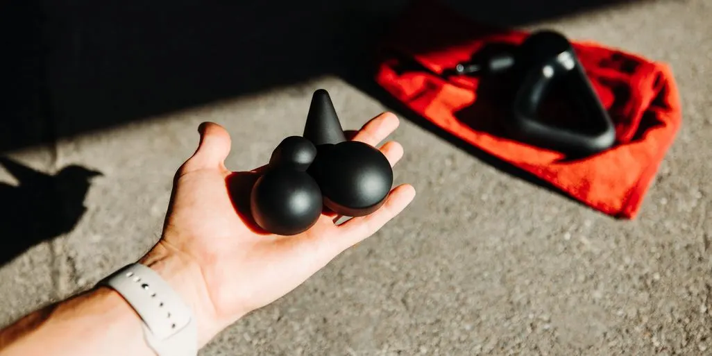 Eine Hand hält verschiedene Aufsätze für eine Massagepistole, im Hintergrund ein rotes Handtuch auf dem die Massagepistole liegt.