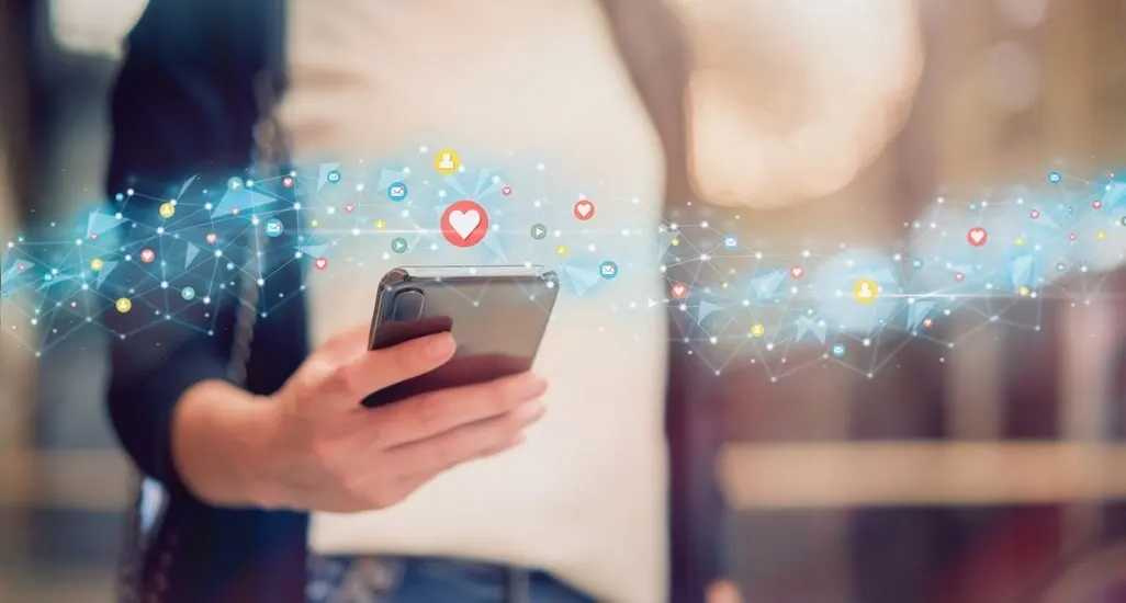 Hand haelt ein Smartphone, darauf schweben Symbole fuer Likes und Herzen