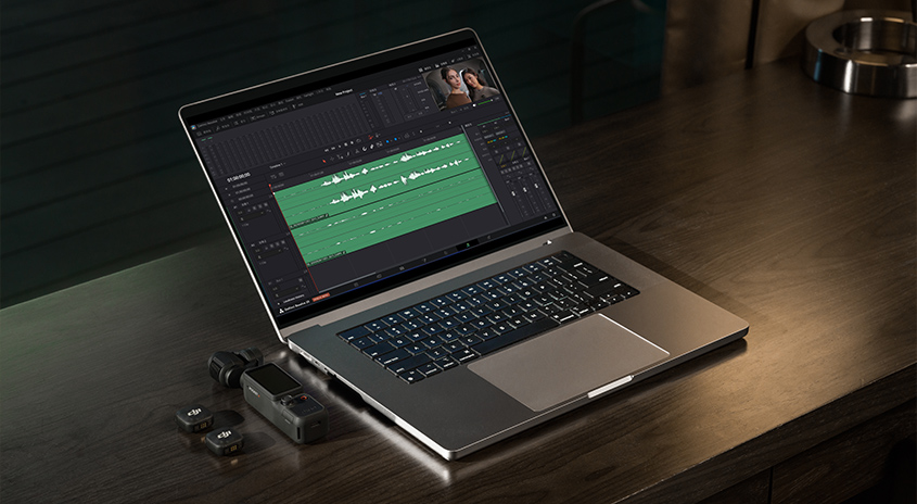 Laptop zeigt Videobearbeitungssoftware mit grüner Audiospur. Daneben professionelle Kamera und Mikrofon auf Holztisch. Kreative Arbeitsstation.