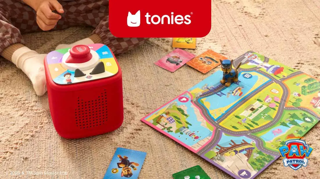 Zwei Kinder sitzen auf einem weißen Sitzsack. Ein Mädchen hält eine rote Toniebox, ein Junge setzt eine rote Figur darauf. Im Hintergrund ein Fenster mit Pflanzen.