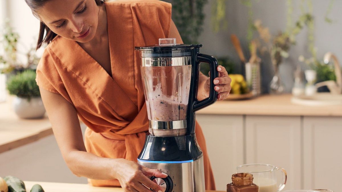 Eine Person in orangefarbenem Kleid bedient einen Standmixer auf einer Küchenarbeitsfläche.