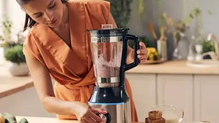 Eine Person in orangefarbenem Kleid bedient einen Standmixer auf einer Küchenarbeitsfläche.
