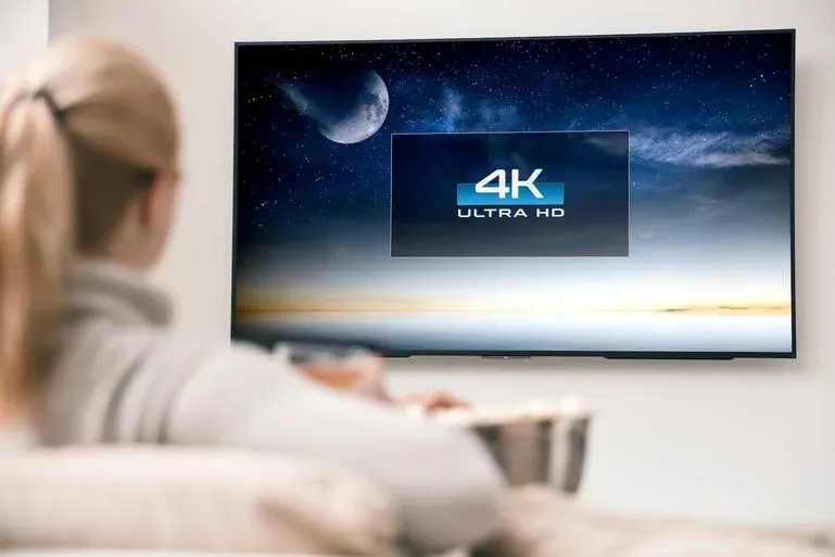 Eine Person sitzt auf einem Sofa, hält eine Schüssel und schaut auf einen Fernseher mit "4K ULTRA HD"-Anzeige und Sternenhimmel.
