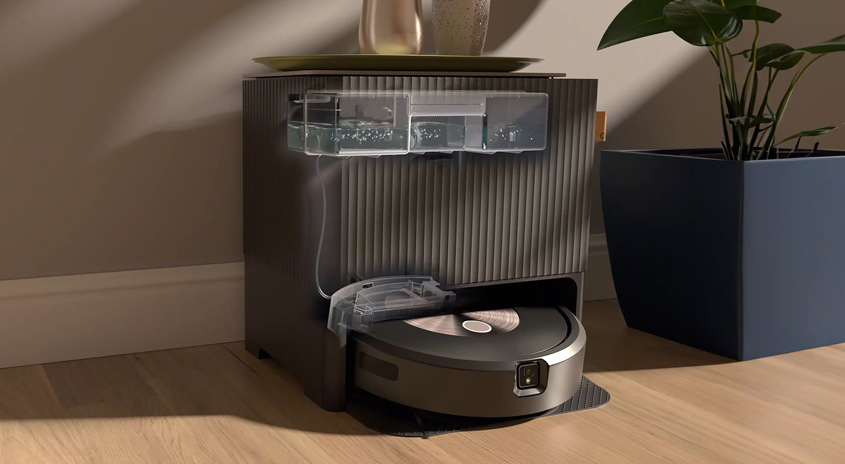 Ein runder Roboterstaubsauger parkt in einer Ladestation mit Wassertanks. Daneben steht eine Topfpflanze auf einem Holzboden.