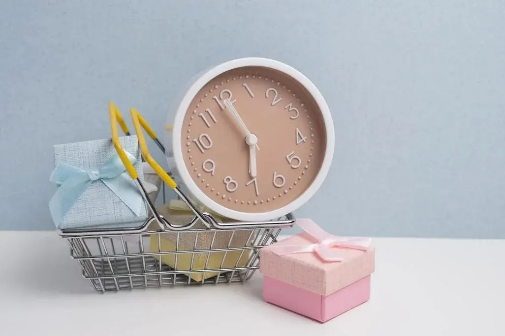 Kleiner Einkaufskorb mit Geschenkboxen, darauf eine runde Pastell-Uhr, daneben eine rosa Geschenkbox mit Schleife.