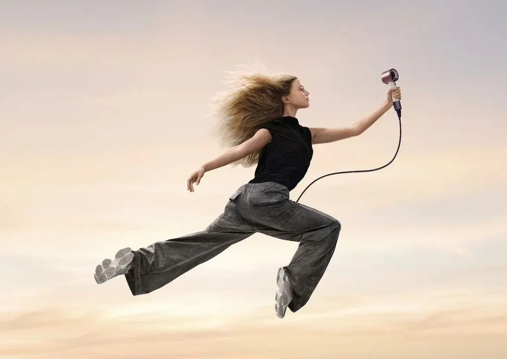 Frau mit wehenden blonden Haaren springt dynamisch in die Luft, trägt schwarzes T-Shirt und graue Jeans, hält rosafarbene In-Ear-Kopfhörer mit schwarzem Kabel in der Hand vor bewölktem Himmel – Bewegung und Energie symbolisierend.