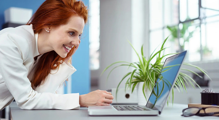 Eine Person in einem weißen Blazer lehnt sich an einen grauen Schreibtisch mit einem Laptop, einer Pflanze, einer Brille und einer schwarzen Schachtel.