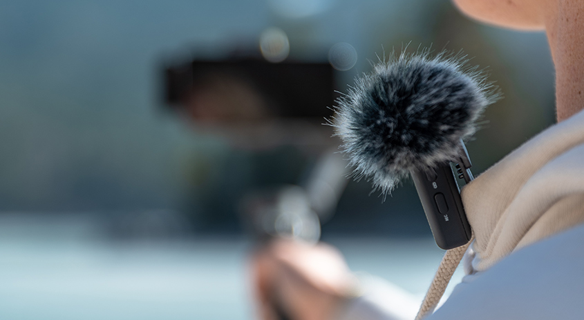 Nahaufnahme eines Mikrofons mit Fellwindschutz, befestigt an einem weißen Kragen, mit verschwommenem Hintergrund.