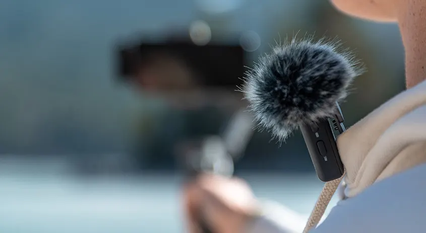 Nahaufnahme eines Mikrofons mit Fellwindschutz, befestigt an einem weißen Kragen, mit verschwommenem Hintergrund.