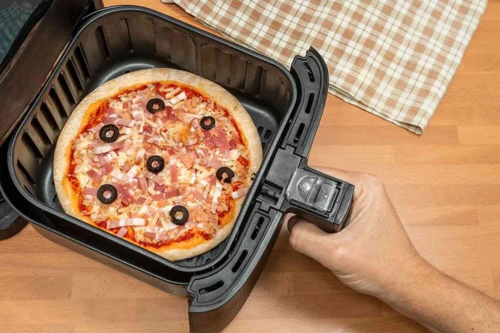 Hand öffnet die Lade der Heißluftfritteuse. Im Korb liegt eine Pizza mit Schinken, Käse und Olivenringen.