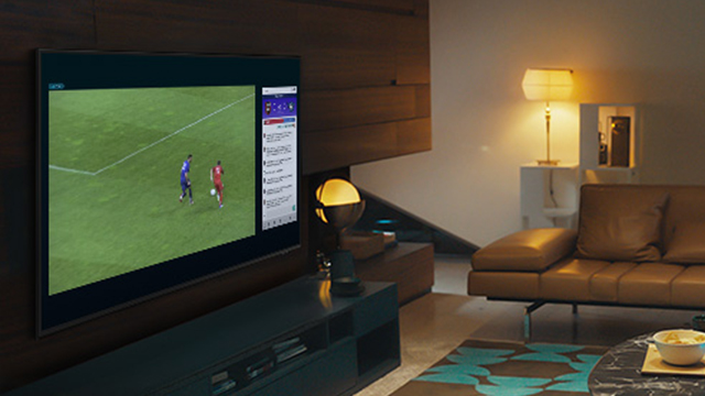 Ein Wohnzimmer zeigt einen Fernseher mit einem Fußballspiel und einer Menüleiste, eine braune Couch und eine Lampe.