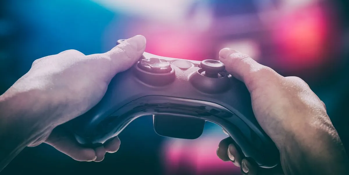 Zwei Hände halten einen schwarzen Gamecontroller vor einem Bildschirm mit buntem Licht im Hintergrund