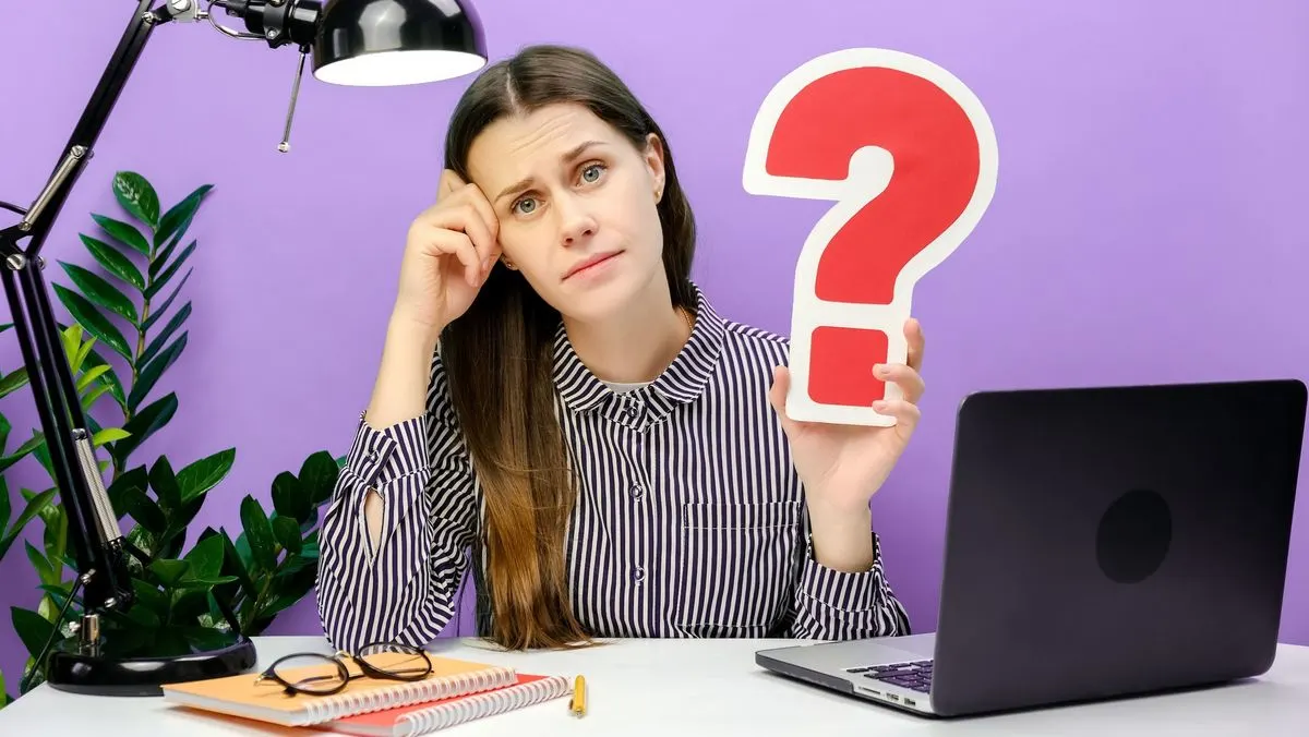 Frau sitzt an einem Schreibtisch mit Laptop, Lampe, Notizbüchern und hält ein großes rotes Fragezeichen aus Pappe hoch