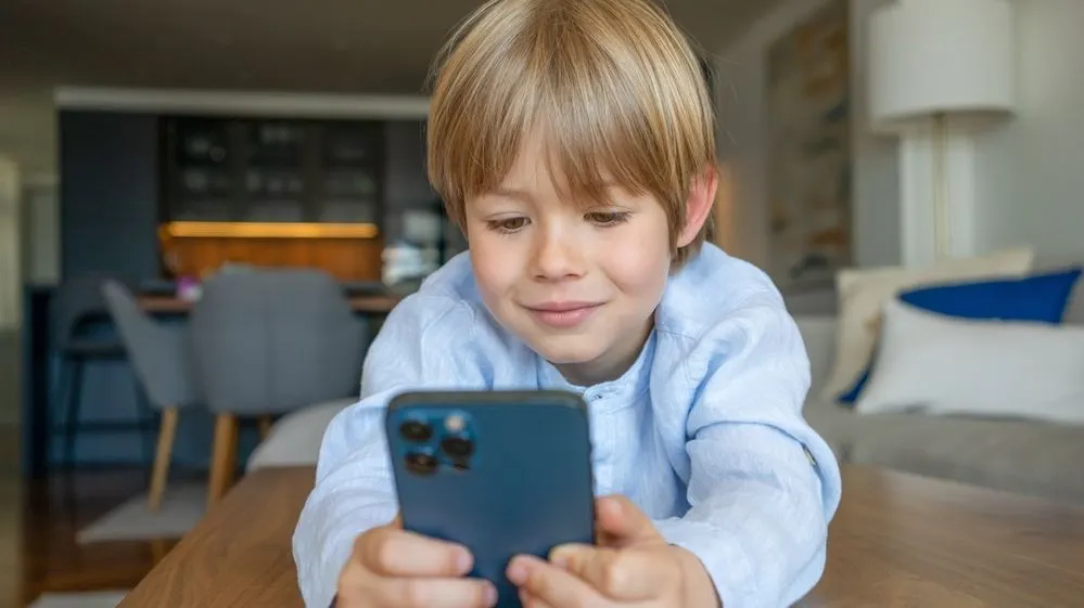 Ein Junge in hellblauem Hemd hält ein dunkelblaues Smartphone, darauf blickend und lächelnd.