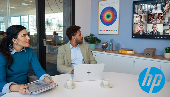 2 Menschen sitzen in einem Besprechungsraum mit einem HP Notebook, an der Wand befindet sich ein Monitor mit zugeschaltenen Personen via MS Teams
