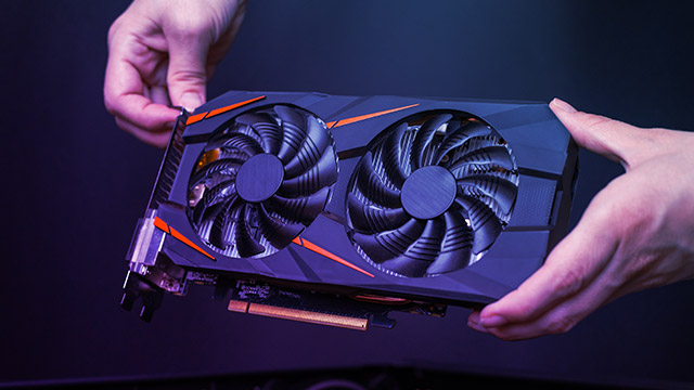 Eine Grafikkarte mit zwei Lüftern wird von zwei Händen vor einem dunklen Hintergrund gehalten.