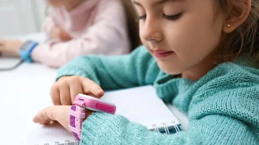 Mädchen sitzt mit einem Schreibblock in der Klasse und bedient ihre Smartwatch für Kinder.