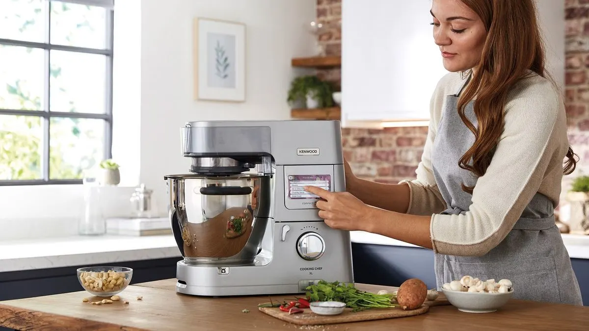 Eine Person in cremefarbenem Pullover und grauer Schürze bedient eine silberne Kenwood Küchenmaschine. Auf der Arbeitsplatte liegen Erdnüsse, Kräuter und Pilze. Heller Küchenhintergrund mit Fenster und Ziegelwand.