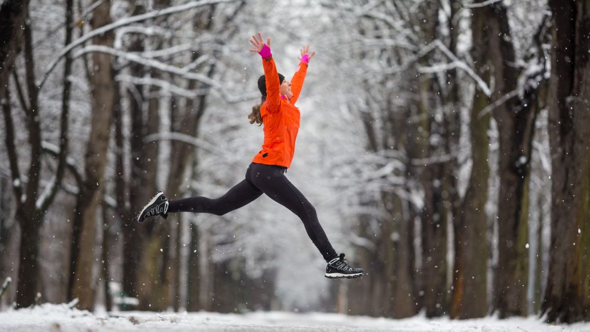 Eine Frau in orangefarbener Jacke springt beim Joggen durch eine verschneite Allee.