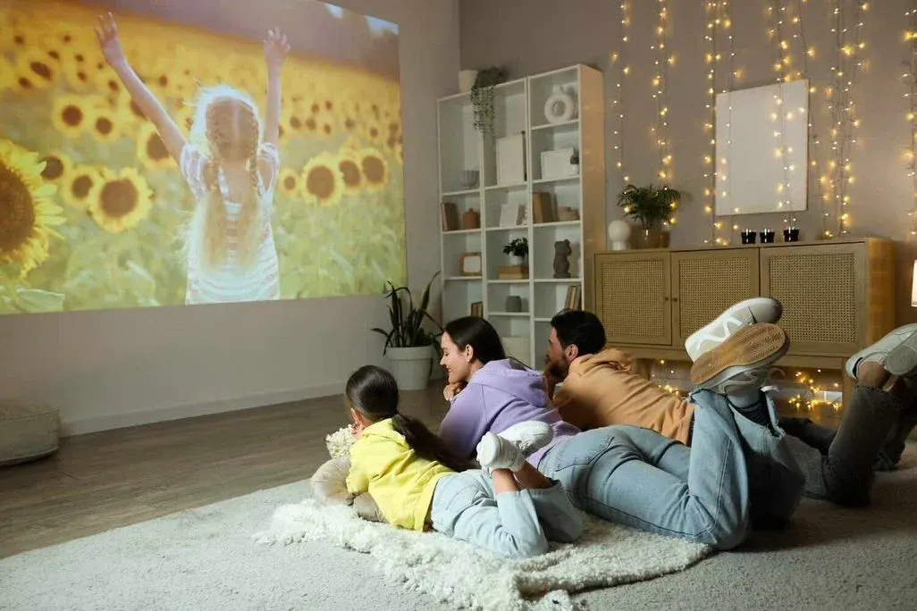Familie liegt gemütlich auf einem Teppich im Wohnzimmer und schaut eine Beamer-Projektion eines Sonnenblumenfelds mit spielendem Kind an der Wand.