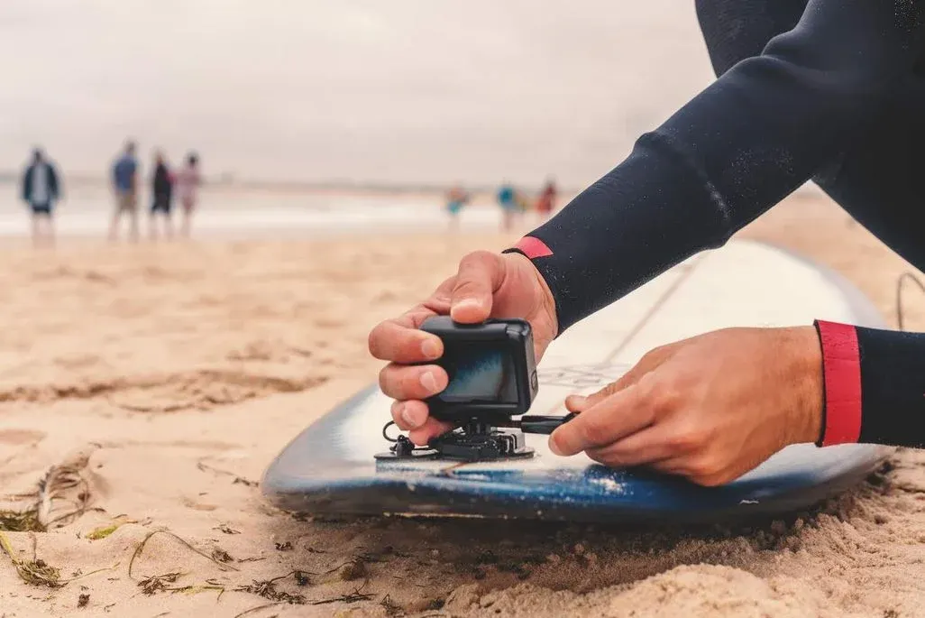 Eine Person im dunkelblauen Neoprenanzug mit roten Ärmelbündchen kniet am Sandstrand und befestigt mit beiden Händen eine kleine schwarze Action-Kamera (vermutlich eine GoPro) auf einer Halterung auf einem blauen Surfboard. Das Surfboard liegt auf dem Sand. Im unscharfen Hintergrund sind mehrere Personen am Strand sowie ein bewölkter Himmel zu erkennen. Die Szene wirkt wie eine Vorbereitung für eine Surf-Session.