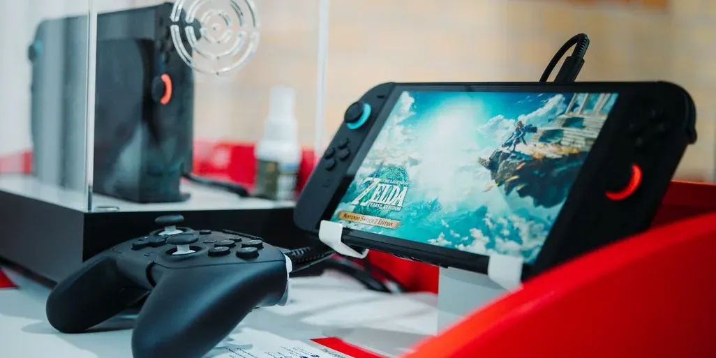 Handheld-Konsole auf Ständer mit Spielbildschirm, daneben ein Gamepad und eine Dockingstation/Box auf einem Tisch.