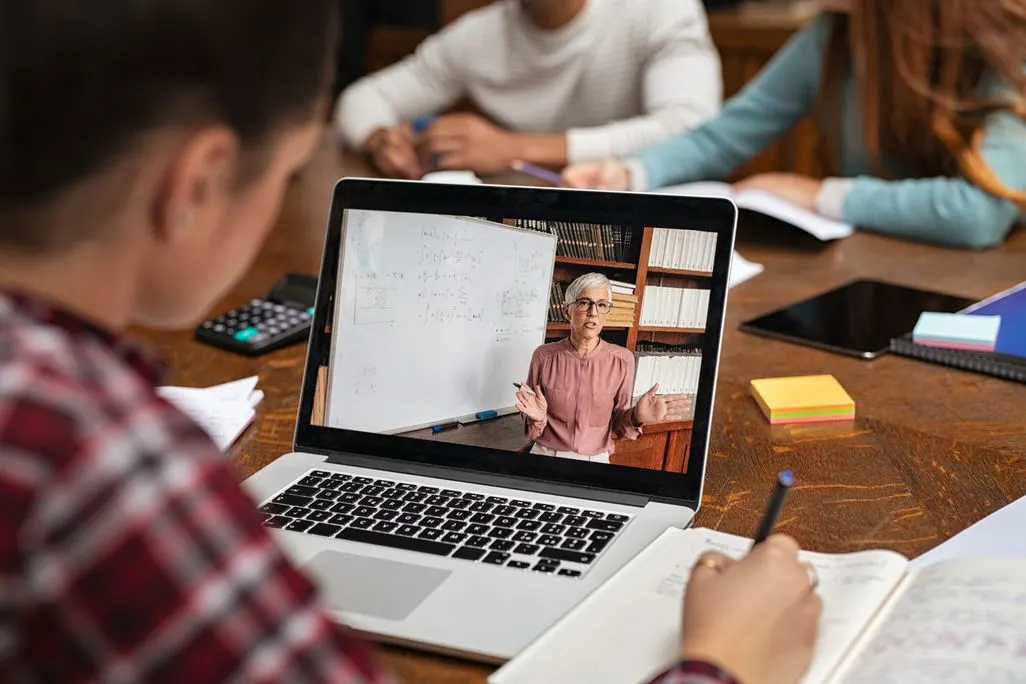 Schüler*in lernt am Laptop von Lehrerin vor Tafel/Bücherregal. Auf Holztisch: Notizbuch, Taschenrechner, bunte Notizblöcke.