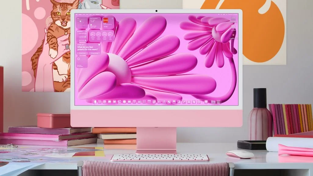 Pinker iMac mit Blumen-Wallpaper, Tastatur, Maus auf einem Schreibtisch mit rosa Büchern, Schachtel, Stiften. Katzen- und abstrakte Kunst an der Wand.