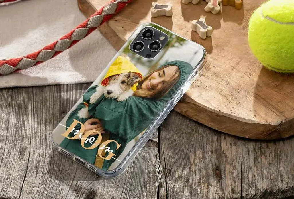 Smartphone mit personalisierter CEWE‑Handyhülle, die ein Foto einer Person in grüner Regenjacke zeigt, die einen kleinen Hund im gelben Regenmantel hält; Gerät liegt auf einer Holzfläche neben Tennisball und Hundespielzeug.