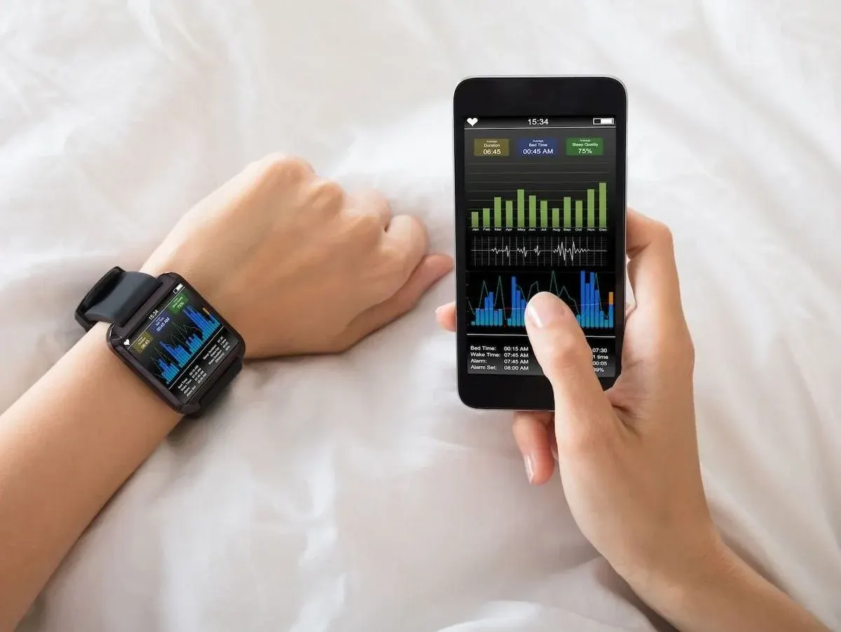 Nahaufnahme von zwei Händen auf weißer Bettdecke; am linken Handgelenk eine Smartwatch und in der rechten Hand ein Smartphone, beide Bildschirme zeigen Balken‑ und Liniendiagramme mit Schlaf‑ und Gesundheitsdaten (z. B. Schlafdauer und Herzfrequenz)