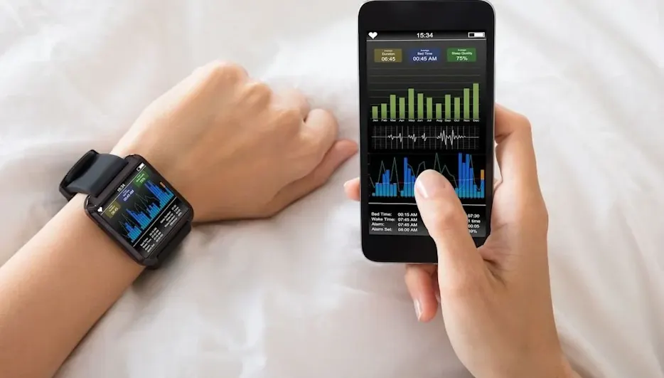 Nahaufnahme von zwei Händen auf weißer Bettdecke; am linken Handgelenk eine Smartwatch und in der rechten Hand ein Smartphone, beide Bildschirme zeigen Balken‑ und Liniendiagramme mit Schlaf‑ und Gesundheitsdaten (z. B. Schlafdauer und Herzfrequenz)