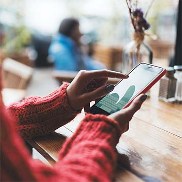 Hände tippen auf ein Smartphone auf einem Holztisch. Im Hintergrund verschwommene Pflanzen und eine Person. Roter Pullover im Vordergrund.