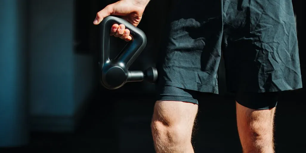 Ein Mann in schwarzen Sportshorts hält eine schwarze Massagepistole in der rechten Hand und massiert seinen Oberschenkel.
